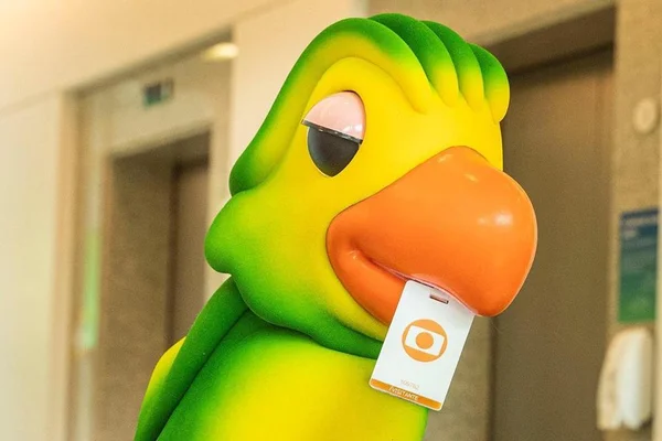 Boneco Louro José, mascote do programa Mais Você, segura crachá da Globo com o bico - Metrópoles