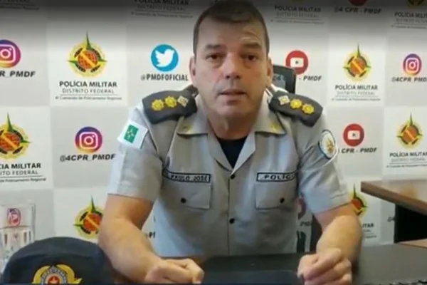 Fotografia colorida de policial militar fardado sentado em cadeira e com braços apoiados em mesa