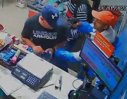Foto colorida mostra assaltante de boné laranja e casaco azul apontando arma para a cabeça de homem com boné da under armour e camiseta da under armour