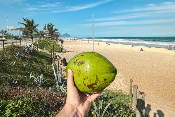 Imagem mostra uma mão segurando um coco verde com canudo em uma praia - Metrópoles