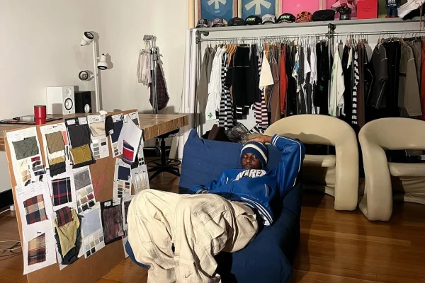 A imagem mostra um homem deitado em um sofá com diversas roupas ao fundo em um arara