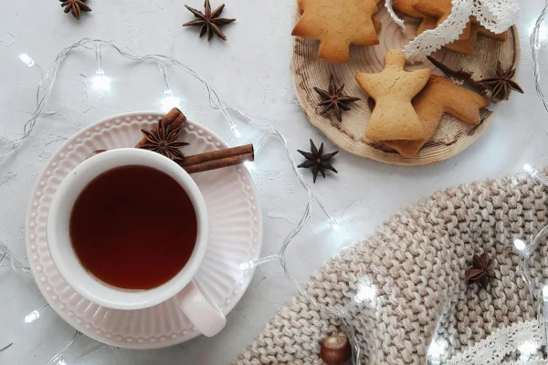 Foto colorida de uma xícara com chá de anis-estrelado, do lado alguns biscoitos e luzinhas decorando
