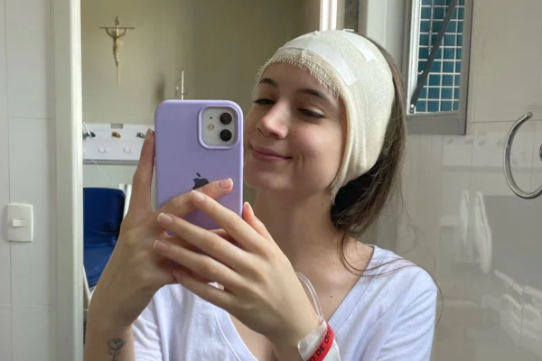 Imagem colorida de jovem segurando celular da capinha roxa enquanto sorri no espelho e tem faixa enrolada na cabeça - Metrópoles