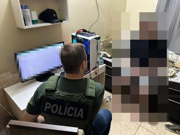 Policial branco, de cabelo curto e claro, usando colete verde, sentado em uma cadeira, de costas para a câmera, em frente a uma mesa com computador com tela borrada. Na frente dele, há a imagem borrada de um adolescente, sentado sobre a cama. Sobre a mesa do computador, há uma prateleira com dois frascos de desodorante e um boné. As paredes e o piso têm cor clara, e a cortina do quarto, à direita, é cinza - Metrópoles