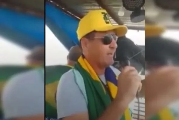 Fotografia colorida de homem com boné amarelo na cabeça e bandeira do brasil em volta do pescoço
