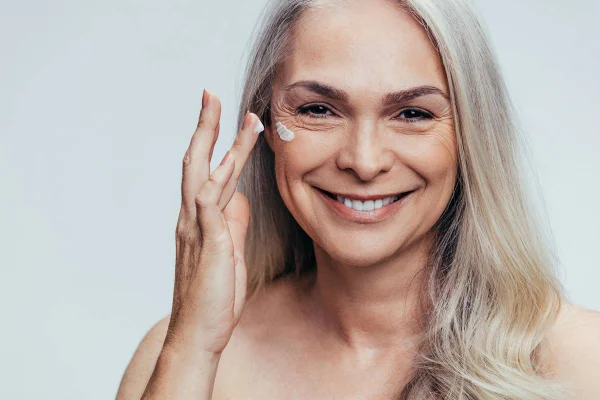 Foto colorida de uma mulher de cabelo grisalho sorrindo enquanto passa creme no rosto com os dedos - Metrópoles