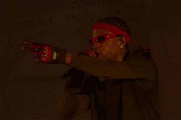 A imagem mostra o rapper Travis Scott durante um show. Com o microfone na mão direita, o artista canta usando bandana e óculos vermelho, junto a u par de luvas na mesma cor.