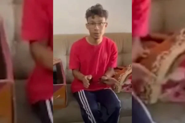 Rapaz branco, de bigode e cavanhaque ralos, de óculos e cabelo curto encaracolado, trajando camiseta vermelha, sentado em sofá, com mãos algemadas para frente - Metrópoles