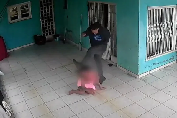 Homem de calça jeans cinza e camiseta de moletom preta da chute em rosto de mulher, de camiseta e saia rosas, caída no chão de garagem de residência - Metrópoles