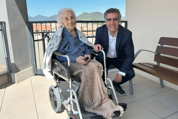 Foto colorida de mulher idosa em cadeira de rodas ao lado de home de paletó - Metrópoles