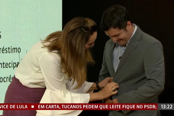 A apresentadora da Globo News Maria Beltrão ajuda convidado que deixou cair o microfone. Na foto, ela ajeita o microfone no terno dele - Metrópoles