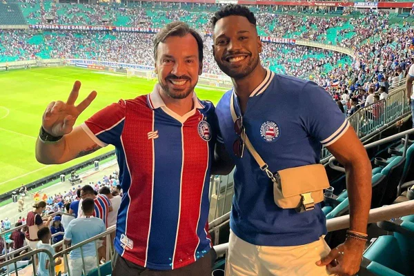 foto colorida de dois homens, um branco de barba e blusa listrada vermelha e azul e um negro de barba rala e blusa azul, posando abraçados - metrópoles