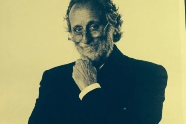 Foto com efeito sépia com fundo amarelado e imagem em preto e branco de homem grisalho, com barba curta. Ele usa óculos, blazer preto e leva uma das mãos ao queixo - Metrópoles