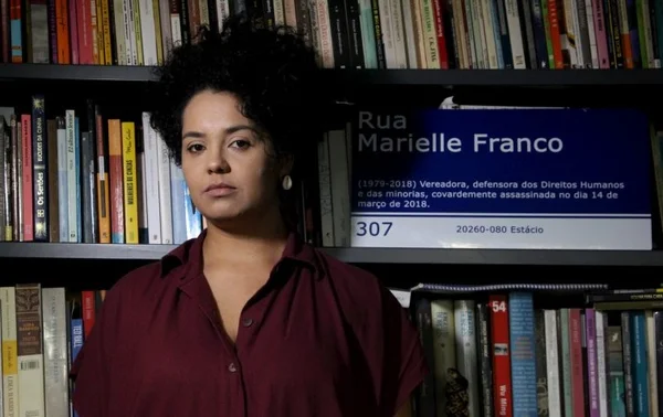A jornalista Bianca Santana posa em frente a uma estante com uma placa de rua que traz o nome da vereadora Marielle Franco