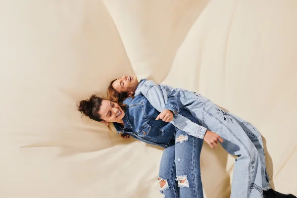 Uma mulher deitada nas costas de outra mulher. Ambas usam jeans, com fundo claro de pano. Campanha de Dia dos Namorados da Renner