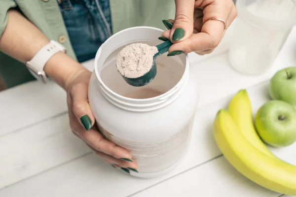 Foto de mulher de calça jeans e camisa segurando colher de medida com porção de proteína de soro de leite em pó acima do frasco de plástico na mesa de madeira branca com coqueteleira, banana e maçã. Processo de fabricação de bebida proteica - Metrópoles