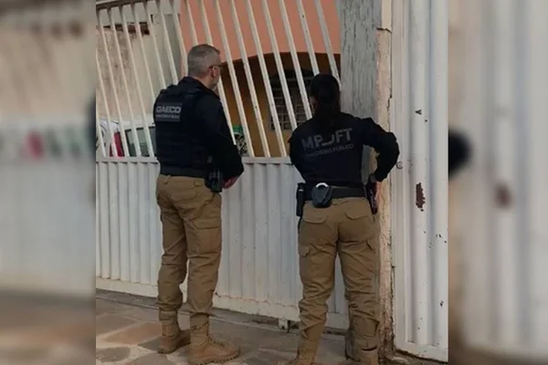 Servidores do MP cumprem mandado de busca e apreensão. Na foto um homem e uma mulher com uniforme preto e calça caqui batem no portão branco