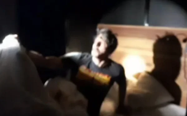 Homem de camiseta preta sentado sobre cama, com luz de lanterna apontada para seu rosto - Metrópoles