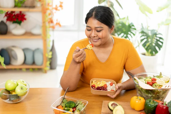 Foto colorida de mulher sentada comendo salada. Ela está em uma mesa com legumes e verduras - Metrópoles