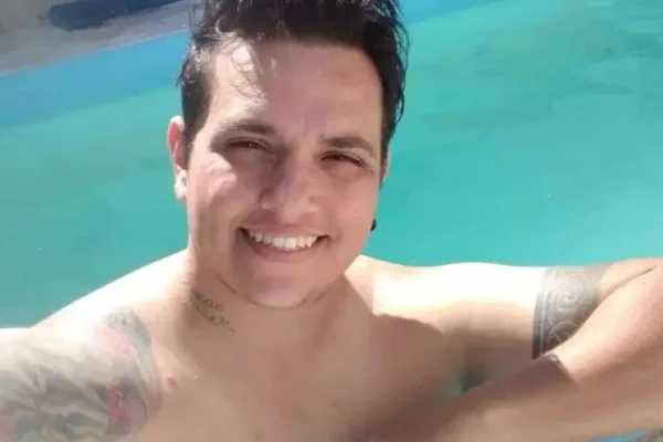 Imagem colorida mostra jovem branco com cabelos escuros curtos. Ele olha para a câmera e sorri mostrando os dentes, O homem está dentro de uma piscina - Metrópoles