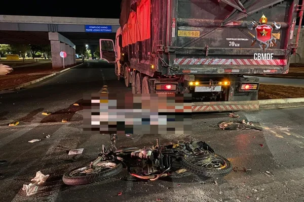 Cena de acidente de trânsito, com moto caída ao chão, atrás de caminhão com lona laranja parado na via; ao fundo, vê-se um viaduto