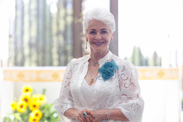 Jane Godoy, uma mulher de 80 anos, cabelos brancos. Está usando um vestido branco com uma flor azul - Metrópoles