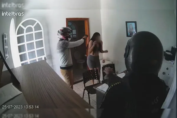 Homem com capacete de moto aponta arma para mulher dentro de uma sala. Outro homem com capacete de moto e outro homem com casaco cinza estão na sala