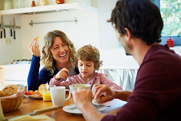 Foto colorida. Família feliz de três pessoas (uma mulher, uma criança e um homem), todos brancos, tomando café da manhã à mesa na cozinha - Metrópoles