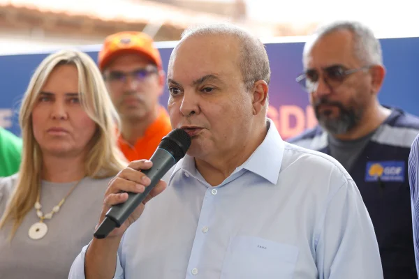 Imagem colorida mostra governador Ibaneis Rocha falando ao microfone. Ele veste camisa social azul-clara. Do lado direito dele, ao fundo, está a vice-governadora Celila Leão. Ela veste blusa cinza e usa um colar dourado. Atrás deles, há dois homens: um vestido com uniforme dos bombeiros, o outro com colete do GDF