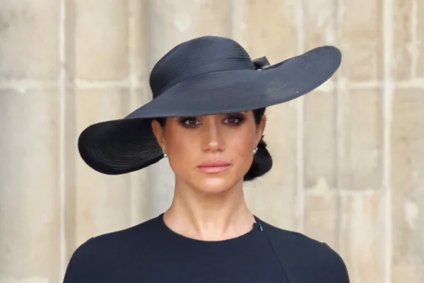 Foto colorida. Na imagem, Meghan Markle, uma mulher negra, está olhando séria para frente. Ela usa um vestido e um chapéu preto - Metrópoles