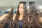 A jovem possui uma pinta bem característica na bochecha direita, próxima ao canto da boca, e cabelos castanhos escuros, longos e cacheados A jovem possui uma pinta bem característica na bochecha direita, próxima ao canto da boca, e cabelos castanhos escuros, longos e cacheados