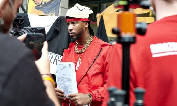 homem negro posa para foto vestindo uma jaqueta vermelha e segurando um papel