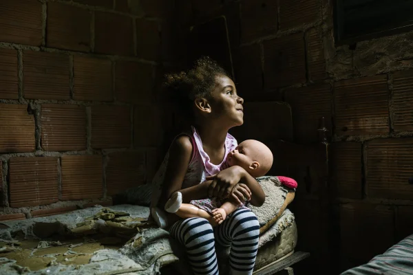 Imagem mostra uma criança sentada em um colchonete em uma casa de tijolos aparentes e segurando uma boneca - Metrópoles
