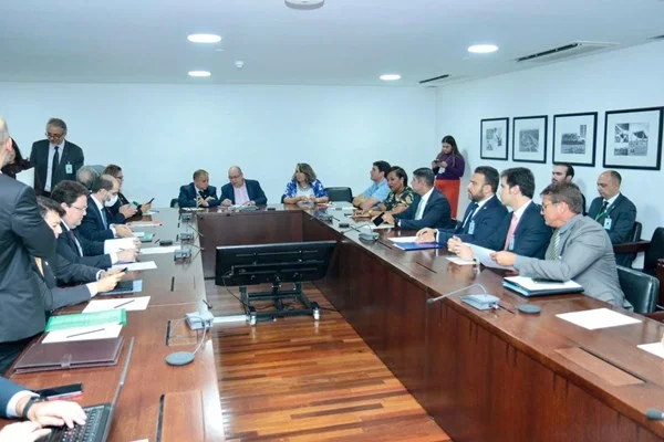 Fotografia colorida mostra grupo de pessoas sentadas em mesa de reunião