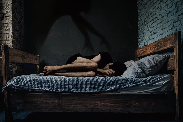 Imagem captura a essência arrepiante da paralisia do sono: uma pessoa encolhida de medo, com uma mão sombria pairando sobre a cabeça, simbolizando o desamparo e o terror da experiência. Metrópoles