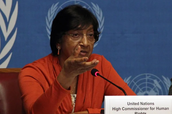 Navi Pillay usa uma blusa laranja e óculos, ela está sentada com o fundo da ONU
