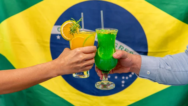 Na foto, duas pessoas brindando com drinques verde e amarelo com a bandeira do Brasil atrás - Metrópoles