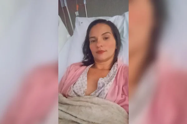 Imagem colorida de mulher branca com casaco rosa deitada em uma cama de hospital - Metrópoles