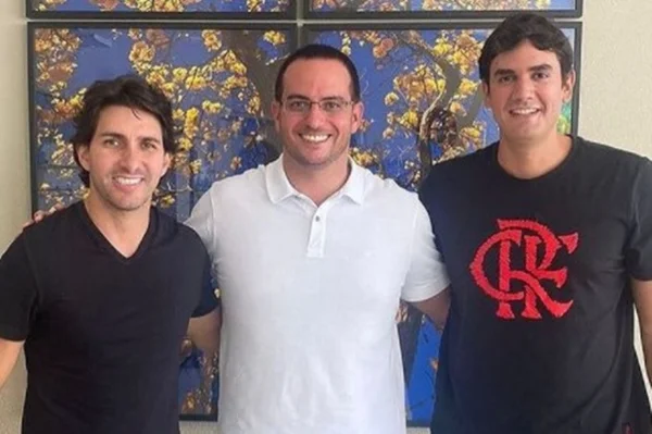 Três homens sorrindo para foto em frente a quadro azul com flores amarelas