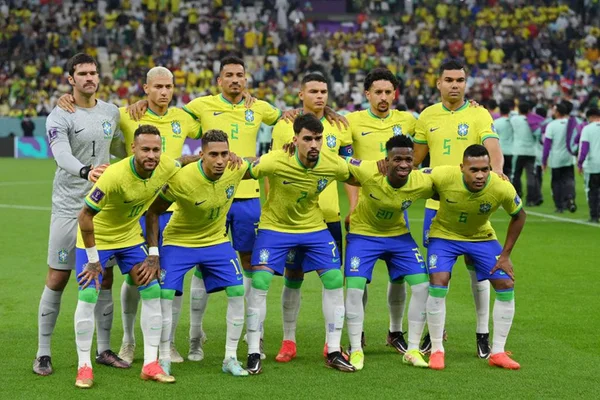 Jogadores da seleção do Brasil em pose de formação para foto em campo, com arquibancada de estádio ao fundo antes de jogo contra a Sérvia na Copa do Mundo do Catar. Eles usam camisa amarela e short azul - Metrópoles