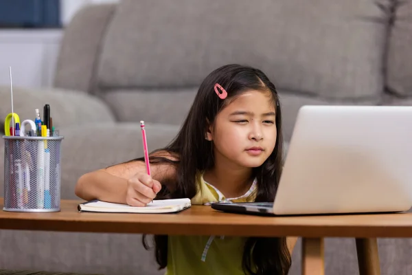 Menina sentada estudando escrevendo e estreitando os olhos para enxergar o computador. Metrópoles