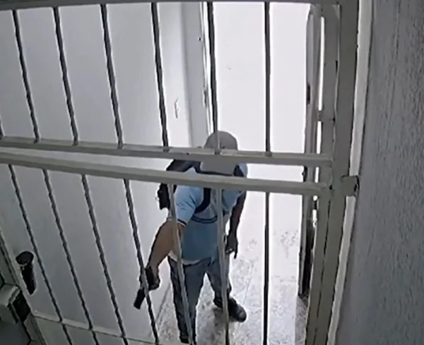 Imagem colorida mostra homem vestido com uma camiseta azul clara com uma arma na mão na frente de um portão com grades. Ele atira em uma mulher na zona leste de São Paulo - Metrópoles