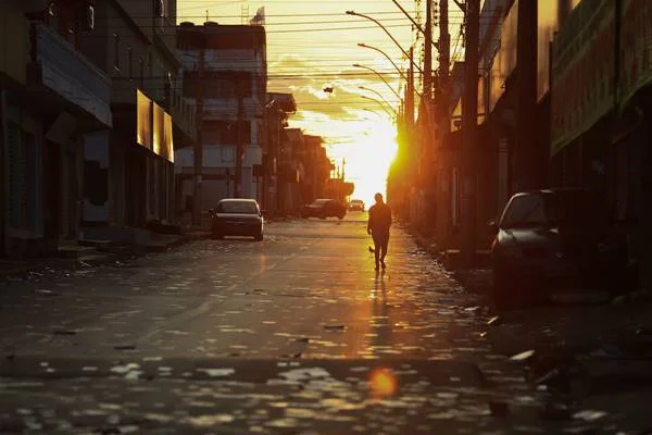 Fotografia colorida de avenida com santinhos jogados no chão, pessoa andando na rua e nascer do sol ao fundo