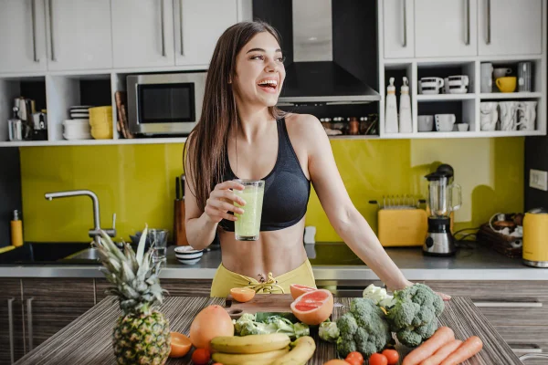 Foto colorida de mulher segurando copo. Ela está em uma cozinha e perto de uma bancada com frutas e legumes - Metrópoles