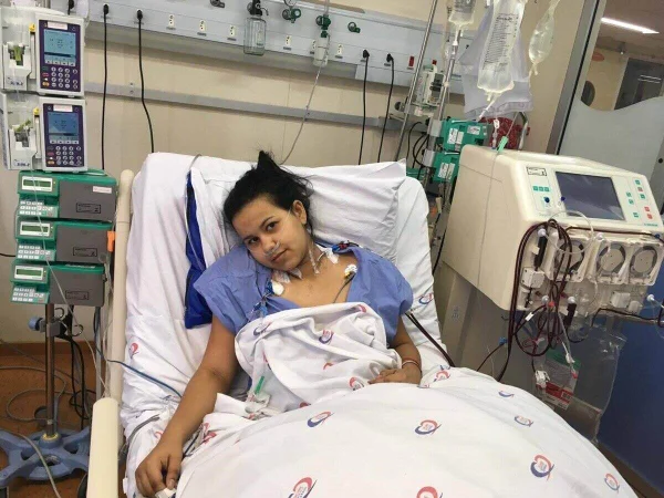 Em foto colorida jovem de camisola hospitalar azul está sobre cama, coberta com lençol brancoe braço direito esticado, no qual está plugado um cateter de soro - Metrópoles
