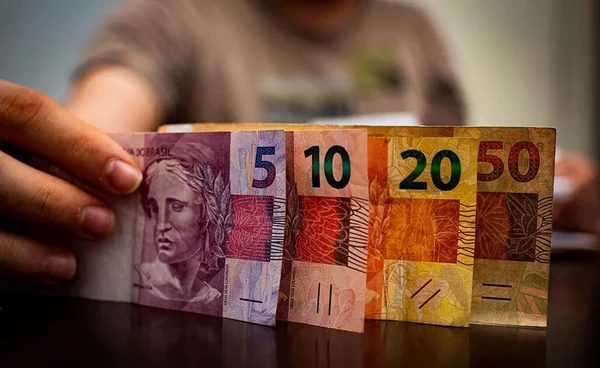 Na imagem colorida, uma mão segura cédulas de cinco, dez, vinte e cinquenta reais