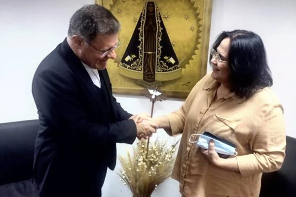 Fotografia colorida de cardeal cumprimentando mulher vestida com roupa bege