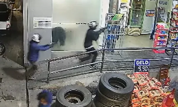 Em foto colorida, criminoso aparece segurando um fuzil com um parceiro logo atrás. Os dois correm na direção da entrada de uma loja de conveniência de um posto de gasolina. Ambos estão de capacete, um deles de blusa azul e outro de blusa preta - Metrópoles