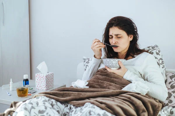 Foto colorida de mulher comendo. Ela está em uma cama e ao lado de frascos de remédio e de uma xícara de chá - Metrópoles