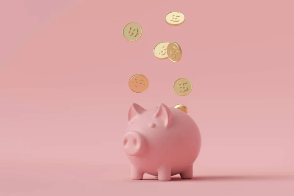 Foto colorida de um cofrinho de porquinho em um fundo cor de rosa com moedas douradas caindo em cima - Metrópoles